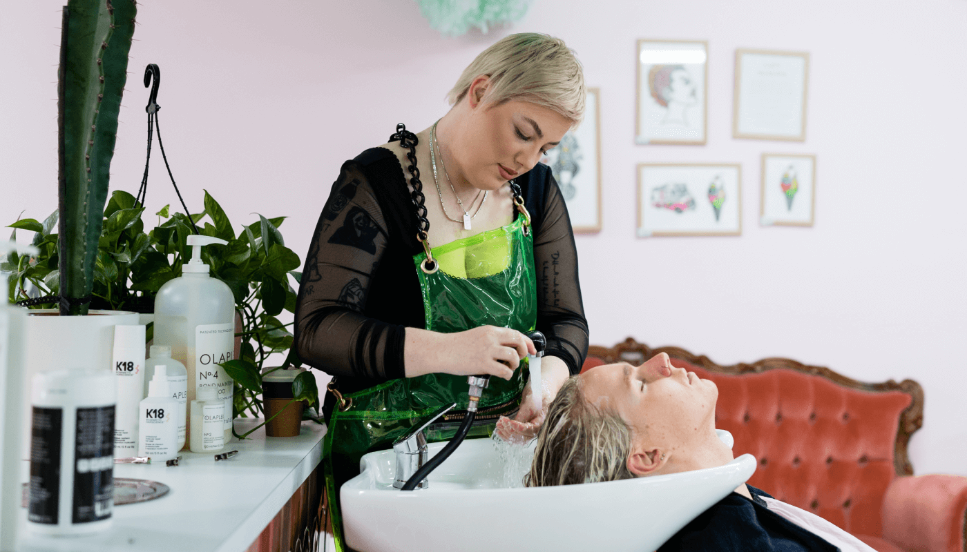 A salon employee is washing a clients hair over a washing basin displaying a variety of professional products they manage using Timely Stock App.